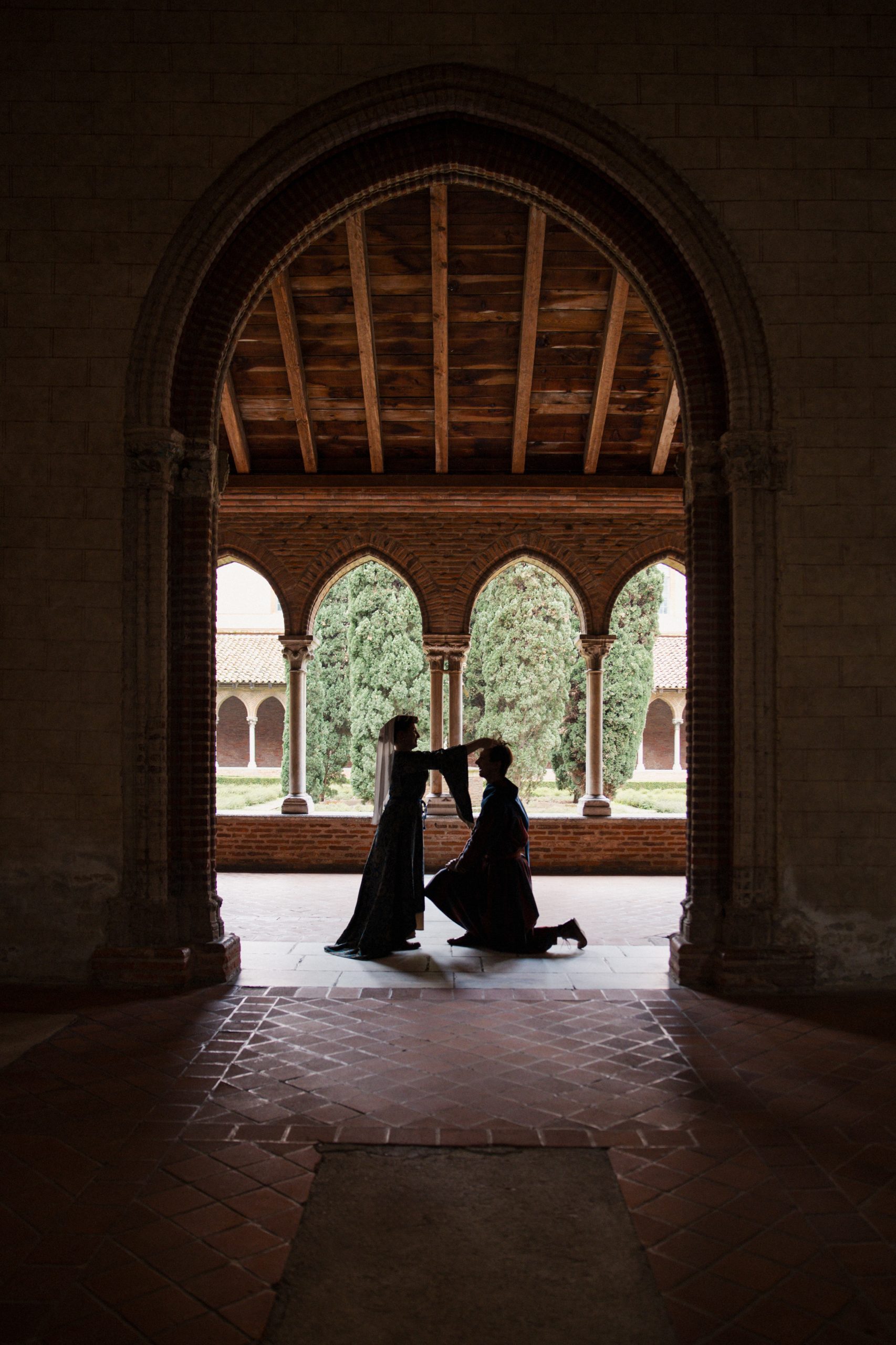 Silhouettes de deux personnes en costumes médiévaux dans une arcade historique : une personne à genoux devant une autre dans un cloître ancien.